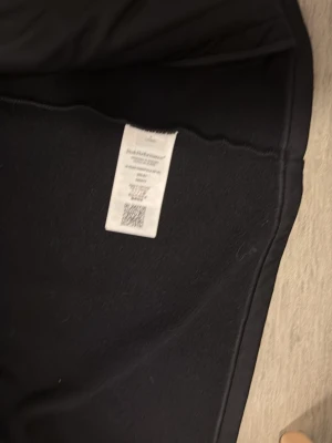 Svart vindjacka från Peak Performance - Snygg svart vindjacka från Peak Performance med huva och dragkedja. Jackan har två fickor med dragkedja och textdetalj på huvan. Tillverkad i ett mjukt och slitstarkt material som passar perfekt för blåsiga dagar.