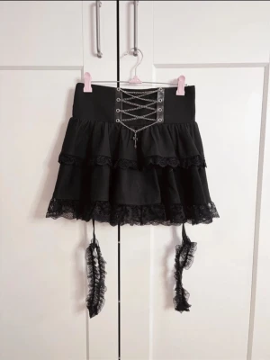 Svart Jirai subcul kjol med snörning och spets - Supersnygg svart kortkjol med volanger, snörning framtill i silverkedja och spetsdetaljer längs kanterna. Kjolen har dekorativa garters som är avtagbara. Från japanska märket Ililil