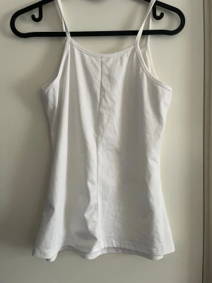 Vit enkel linnetopp - Säljer en vit linnetopp med smala axelband och rak passform. Toppen är ärmlös och gjord i mjuk bomull, perfekt för varma dagar eller som lager under en skjorta. Enkel och stilren design som passar till det mesta.