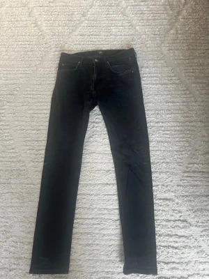 Svarta slim jeans från Lee - Snygga svarta jeans från Lee med klassisk femficksmodell och smal passform. Jeansen har silverfärgade knappar och detaljer samt diskreta slitningar för en cool look. Perfekta till vardagsoutfiten och lätta att matcha. Storlek w30, L 32. Passar om du är runt 170