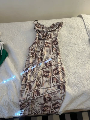 Klänning med dollartryck och öppen rygg - Unik långklänning med dollartryck i beige och bruna toner. Klänningen har smala axelband, omlott-detalj upptill och öppen rygg med knytband. Materialet känns mjukt och stretchigt, troligen polyester. Perfekt för dig som vill sticka ut!