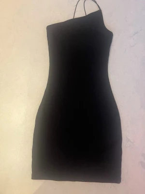 Svart oneshoulder kort klänning - Säljer en svart, tight och ribbad kort klänning med oneshoulder-design och smal axelrem. Klänningen har en enkel och stilren look som passar perfekt till dig som gillar minimalistisk stil. Inte anstånd så mycket