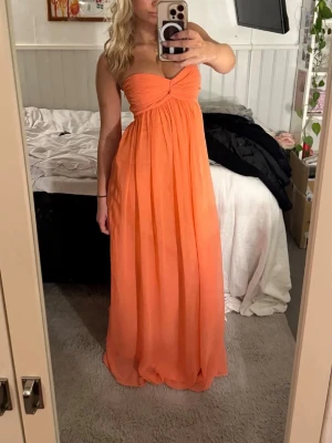 Rosa/orange långklänning - Snygg axelbandslös långklänning i en stark orange färg. Klänningen har en twistad detalj vid bysten och ett luftigt, fallande tyg som ger en elegant siluett. Perfekt för sommarens alla tillfällen. Materialet känns lätt och svalt.