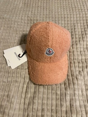 Rosa teddykeps från Moncler - Snygg rosa keps från Moncler i mjukt teddy-material. Klassisk böjd skärm och broderad Moncler-logga framtill. Perfekt accessoar för dig som vill sticka ut med en trendig och mysig look.