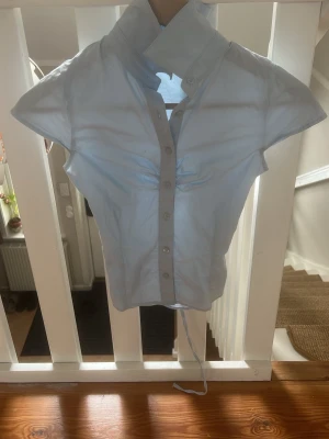 Ljusblå skjorta med snörning - Snygg ljusblå skjorta med korta ärmar och markerad krage. Skjortan har knappar framtill och snörning i midjan för en cool, justerbar passform. Tillverkad i bomull, perfekt för dig som gillar stilrena detaljer.