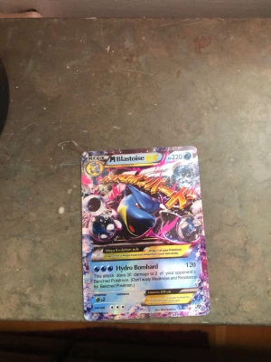 Pokémonkort Mega Blastoise EX - Säljer ett färgstarkt samlarkort från Pokémon, Mega Blastoise EX #220. Kortet har en glansig yta med blå, rosa och gula detaljer och visar Blastoise i en actionfylld pose. Perfekt för dig som vill utöka din Pokémonkort-samling eller spela TCG. Kortet är fejk så det får ett billigt pris.
