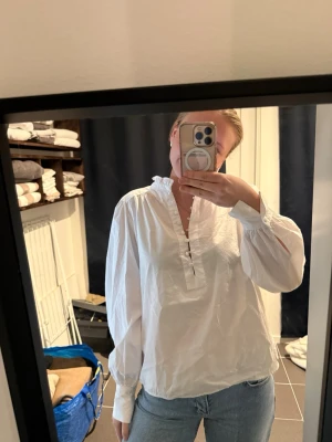 Vit blus med volangkrage - Säljer en vit blus med volangkrage och knappar framtill. Blusen har lång ärm med knapp vid ärmslut och är gjord i ett tunt, luftigt material. Perfekt att styla med jeans eller kjol för en trendig look.