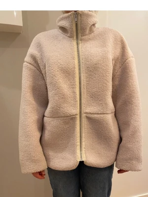 Beige teddyjacka från H&M storlek s - Mysig beige teddyjacka från H&M med dragkedja framtill och mjukt, fluffigt material. Jackan har avslappnad passform, långa ärmar och diskreta sömdetaljer. Perfekt för att hålla värmen under kyliga dagar.
