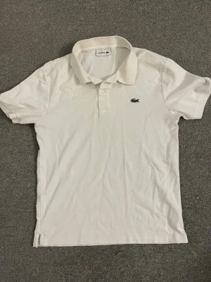 Vit pikétröja från Lacoste - Klassisk vit pikétröja från Lacoste med den ikoniska krokodilloggan på bröstet. Tröjan har krage, korta ärmar och knappar vid halsen. Tillverkad i bomull för en bekväm och luftig känsla.