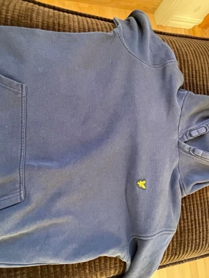 Blå hoodie från Lyle & Scott Junior - Mysig blå hoodie från Lyle & Scott Junior med klassisk gul logga på bröstet. Hoodien har en stor känguruficka framtill och mjuk huva. Perfekt för chill dagar och enkel att matcha med jeans.