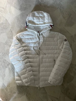 Vit dunjacka Moncler - Snygg vit dunjacka från Moncler med quiltad design. den har en oljefläck på insidan och 2 för täckta hål på baksidan (se på bild)