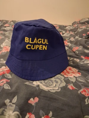 Blå bucket hat Blågul Cupen - Snygg blå bucket hat med gul text 'BLÅGUL CUPEN' broderat framtill. Klassisk form med bred brätte och sydda sömmar. Tillverkad i ett lätt tyg, perfekt för att lyfta din stil på sommaren eller festivalen.