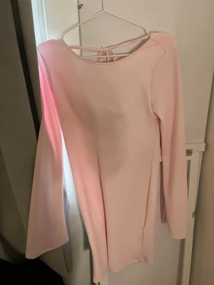 Ljusrosa långärmad klänning med knyt - Söt ljusrosa klänning med långa ärmar och snygg öppen rygg med knytband. Klänningen har en enkel, rak passform och är tillverkad i ett mjukt material som känns bekvämt mot huden. Perfekt för dig som gillar stilrena och feminina plagg.