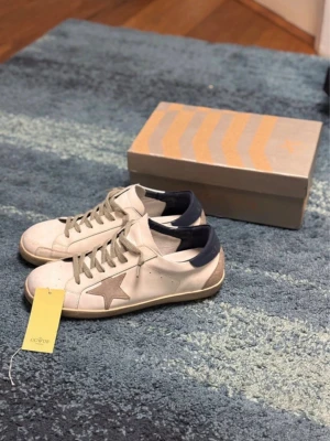 Golden Goose vita sneakers med stjärna - Golden Goose sneakers i vitt läder med grå stjärndetalj på sidan, grå skosnören och mörkblå häl. Klassisk låg modell med slitna detaljer och beige sula. Perfekta för dig som gillar streetstyle och unika detaljer. Jätte fina skor och för mer info skriv gärna!