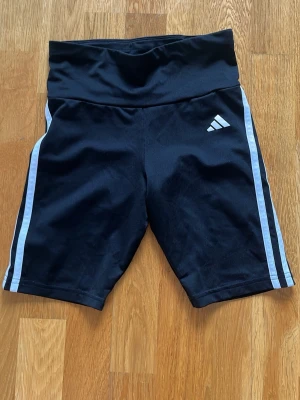 Svarta cykelshorts från Adidas - Snygga svarta cykelshorts från Adidas med vita ränder längs sidorna och Adidas-logga på låret. Shortsen har bred midjeresår för extra komfort och är tillverkade i ett stretchigt funktionsmaterial som passar perfekt till träning eller sport.