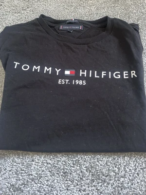 Svart Tommy Hilfiger t-shirt - Snygg svart t-shirt från Tommy Hilfiger med klassisk logga och texten 'EST. 1985' på bröstet. T-shirten har rund hals och korta ärmar. Tillverkad i mjuk bomull som känns skön mot huden. Perfekt för en stilren och avslappnad look.