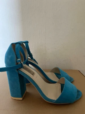 Blåa chunky klackar med rem 10 cm - Snygga blå pumps med bred klack och öppen tå. Skorna har en justerbar ankelrem och är tillverkade i mockaimitation. Perfekta för dig som vill ha både stil och komfort. Märket är Shoex.