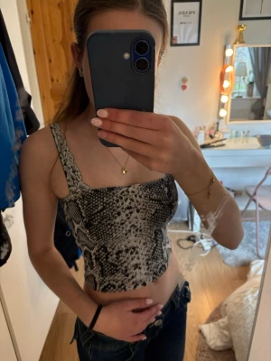 Grå ormmönstrad croppad linnetopp - Säljer en croppad linnetopp med grått ormmönster och spetsdetaljer vid axelbanden. Toppen har en tight passform och är ärmlös, perfekt för dig som vill sticka ut lite extra. Supersnygg till både jeans och kjol!
