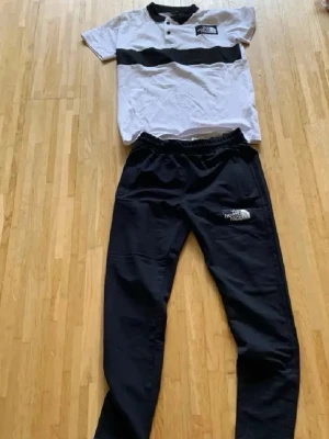 The North Face svartvitt set - Snyggt set från The North Face med en vit t-shirt med svart rand och knappar vid halsen samt svarta byxor med resår i midjan. Båda plaggen har tydlig logga framtill. Perfekt för en avslappnad och sportig stil.
