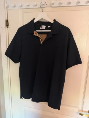 Mörkblå pikétröja från Burberry - Snygg mörkblå pikétröja från Burberry i klassisk modell med kort ärm och krage. Ikoniskt Burberry-rutigt tyg syns på insidan av knäppningen. Perfekt för en stilren och avslappnad look.