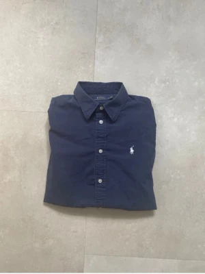 Mörkblå skjorta från Polo Ralph Lauren - Klassisk mörkblå skjorta från Polo Ralph Lauren med vit broderad logga på bröstet. Skjortan har knappar framtill och en stilren krage. Perfekt för en clean och tidlös look.