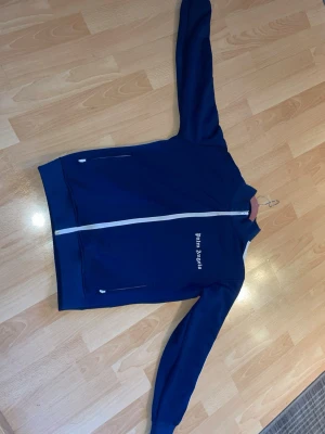 Blå ziptröja från Palm Angels - Snygg blå ziptröja från Palm Angels med vit dragkedja och vita ränder längs ärmarna. Tröjan har två fickor med dragkedja och logga tryckt på bröstet. Perfekt för en sportig och trendig look.
