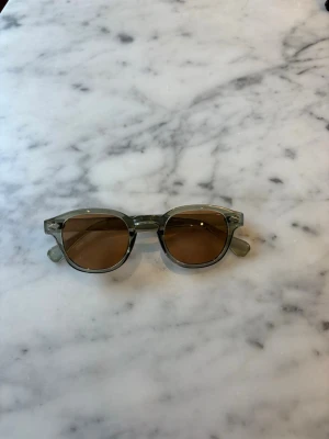 S&T 003 Green solglasögon  - Snygga solglasögon med transparent grön båge och runda bruna glas. Klassisk och trendig modell som passar perfekt till sommaren. Bågarna är i syntetmaterial och har en stilren design utan extra detaljer.