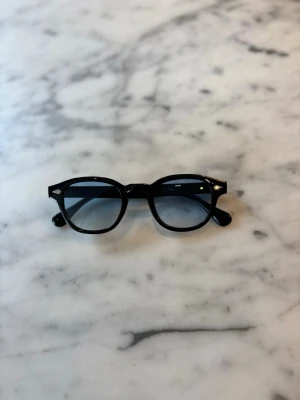 S&T 003 Black solglasögon  - Snygga solglasögon med svart båge och runda blåtonade glas. Klassisk och trendig design som passar till många stilar. Bågarna är i plast och har små silvriga detaljer vid sidorna. Perfekta för soliga dagar.