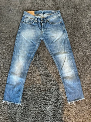 Dondup george jeans - Säljer ett par blå jeans med snyggt slitna detaljer och råa, fransiga benslut. Jeansen har klassisk femficksmodell och en ljus tvätt med markerade slitningar på framsidan. Perfekta för en avslappnad och trendig look.