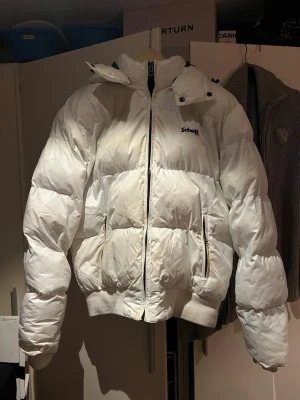 Vit pufferjacka från Schott - Snygg vit pufferjacka från Schott med hög krage, avtagbar huva och dragkedja framtill. Jackan har två sidofickor med dragkedja och ribbade muddar vid ärmslut och midja. Skriv gärna om ni har några frågor eller funderingar :)