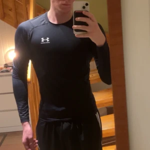 Under armour compression  - Säljer en svart långärmad träningströja från Under Armour med vit logga på bröstet. Tröjan har tight passform och är tillverkad i ett stretchigt funktionsmaterial som andas, perfekt för träning och sport.