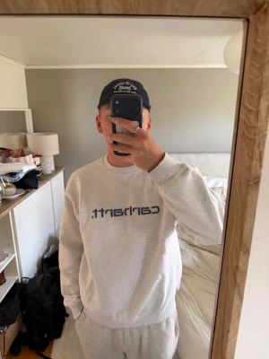 Grå Carhartt sweatshirt - Snygg grå sweatshirt från Carhartt med mörkblå logga tryckt på bröstet. Tröjan har rund halsringning, ribbade muddar och avslappnad passform. Perfekt för en chill och stilren look.