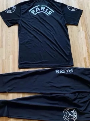 Svart Paris set med tryck - Snyggt svart set med t-shirt och byxor från Paris Saint-Germain. Båda plaggen har vita tryck med texten 'PARIS' och PSG-logga på ärmar och ben. Perfekt för dig som gillar sportig och trendig stil.