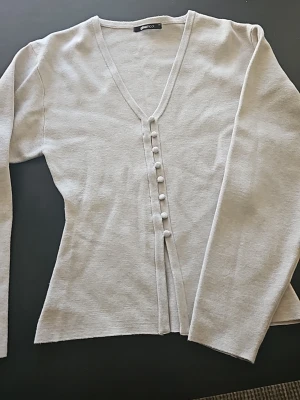 Ljusbeige kofta från Gina Tricot - Snygg ljusbeige kofta från Gina Tricot med v-ringning och dekorativa knappar framtill. Långärmad och i mjukt, tunt stickat material som passar perfekt till lager-på-lager. Enkel och stilren design som är lätt att matcha.