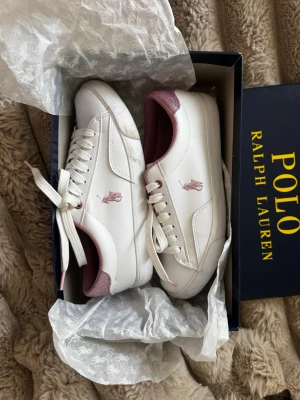Vita Polo Ralph Lauren sneakers - Använd väldigt få gånger. säljer då de var för små. Snygga vita sneakers från Polo Ralph Lauren med rosa detaljer och klassisk logga på sidan. Skorna har lågt skaft, vita snören och rosa insida samt hälparti. Tillverkade i slätt läderliknande material och passar perfekt till en clean och stilren look.