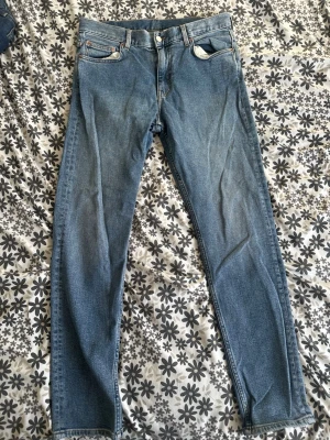 Blå raka jeans med fem fickor - Säljer ett par klassiska blå jeans med rak passform och fem fickor. Jeansen har normal midja, knappgylf och subtila slitningar för en avslappnad look. Materialet är denim i bomull. Perfekta till en casual outfit.