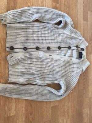 Beige stickad kofta med knappar - Snygg beige stickad kofta med grov struktur och långa ärmar. Koftan har flera mörka knappar framtill och ribbade muddar vid ärmslut och nederkant. 