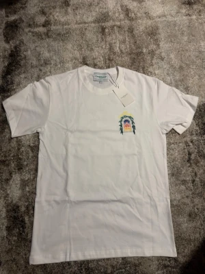 Vit t-shirt från Casablanca - Snygg vit t-shirt från Casablanca med ett färgglatt motiv på bröstet som föreställer ett fönster med blad och texten 'Casablanca'. Klassisk passform och rund hals. Perfekt för dig som gillar stilrena plagg med unika detaljer.