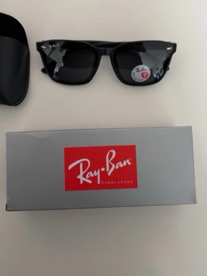 Svarta Ray-Ban Wayfarer solglasögon - Klassiska svarta Ray-Ban Wayfarer solglasögon med polariserade linser. Snygg, tidlös design med rektangulär form och diskret Ray-Ban-logga på bågen. Kommer med originalfodral och box. Perfekta för att lyfta din stil i solen.