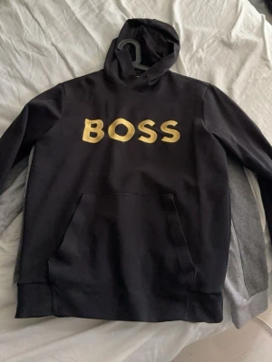 Svart hoodie från BOSS med gul text - Svart hoodie från BOSS med stor gul logga på bröstet. Klassisk modell med huva och känguruficka framtill. Mjuk bomullsblandning som känns skön mot huden. Perfekt för en avslappnad och trendig stil.