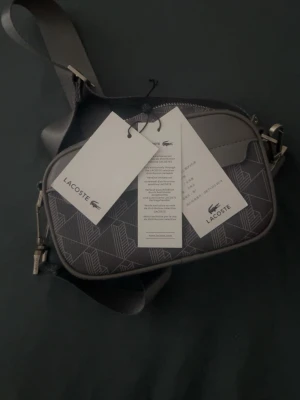 Grå axelväska från Lacoste - Snygg grå axelväska från Lacoste med justerbar axelrem och geometriskt mönster på framsidan. Väskan har en praktisk dragkedja och en extra ficka framtill. Tillverkad i slitstarkt syntetmaterial och har diskret Lacoste-logga.