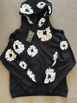 Svart hoodie med vita pufftryck - Cool svart hoodie från Daily Paper med unika vita pufftryck i form av blommor över hela tröjan och huvan. Hoodien har klassisk känguruficka och ribbade muddar. Materialet är mjukt bomullstyg och huva med dragsko ger en avslappnad streetwear-look.