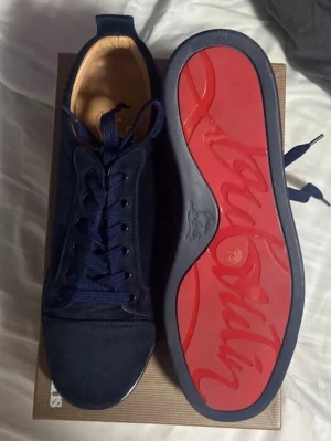  Christian Louboutin  - Snygga mörkblå sneakers från Christian Louboutin med klassisk röd sula och diskret logga undertill. Skorna har ovandel i mocka och matchande blå skosnören. Perfekta för dig som vill sticka ut med exklusiv stil och detaljer.