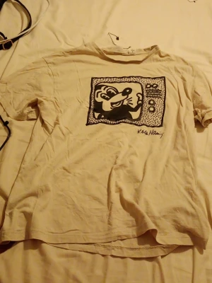 Vit t-shirt Keith Haring x H&M - Vit t-shirt från H&M med coolt svart tryck av Keith Haring på bröstet. Klassisk passform med korta ärmar och rund halsringning. Perfekt för dig som gillar streetart och vill sticka ut med en unik design.