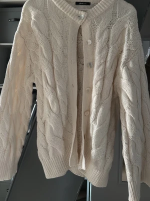 Vit kabelstickad kofta Gina Tricot - Kabelstickad kofta i mjuk vit bomull från Gina Tricot. Koftan har rund halsringning, stora knappar framtill och ribbade muddar vid ärmslut och nederkant. 