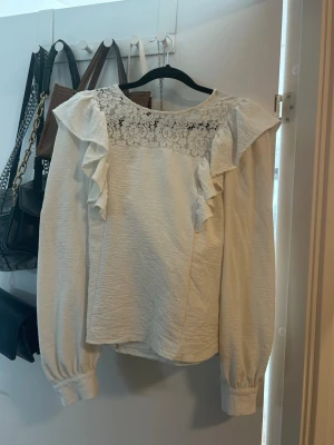 Vit blus med spets och volanger - Söt vit blus med långärmad design, spetsdetalj över bröstet och volanger längs axlarna. Blusen har rund halsringning och puffiga ärmslut med knappar. Perfekt för en trendig och romantisk look.