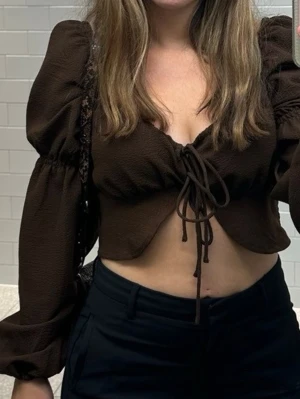 Brun croppad blus med puffärm - Trendig brun blus med croppad passform och långa puffärmar. Toppen har snygg snörning framtill och v-ringning som ger en cool look. Perfekt för dig som vill sticka ut med en unik topp i garderoben.