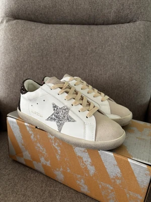 Vita Golden Goose sneakers med glitterstjärna - Snygga vita sneakers från Golden Goose med en glittrig silverstjärna på sidan och beige mockadetaljer vid tån. Skorna har beige snören och svartglittrig häl. Perfekta för dig som vill sticka ut med en cool och trendig look.