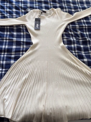 Vit ribbad klänning från FBSister - Supersnygg vit ribbad klänning från FBSister med markerad midja och utsvängd kjol. Klänningen har lång ärm och ett stretchigt material som ger en smickrande passform. Perfekt för dig som vill ha en stilren och trendig look.