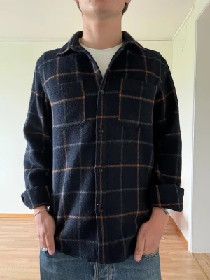 Mörkblå rutig overshirt från A.P.C - Snygg mörkblå overshirt med rutigt mönster i brunt och vitt. Skjortan har två bröstfickor, knäppning framtill och en avslappnad passform. Perfekt att slänga över en t-shirt för en chill look. Materialet känns mjukt och lite tjockare, vilket ger en känsla av en flanell ungefär.